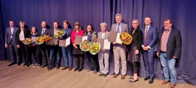 Image shows a row of people in front of a blue curtain background. Some are holding floral bouquets and certificates. These are the recipients of the DGM Awards.