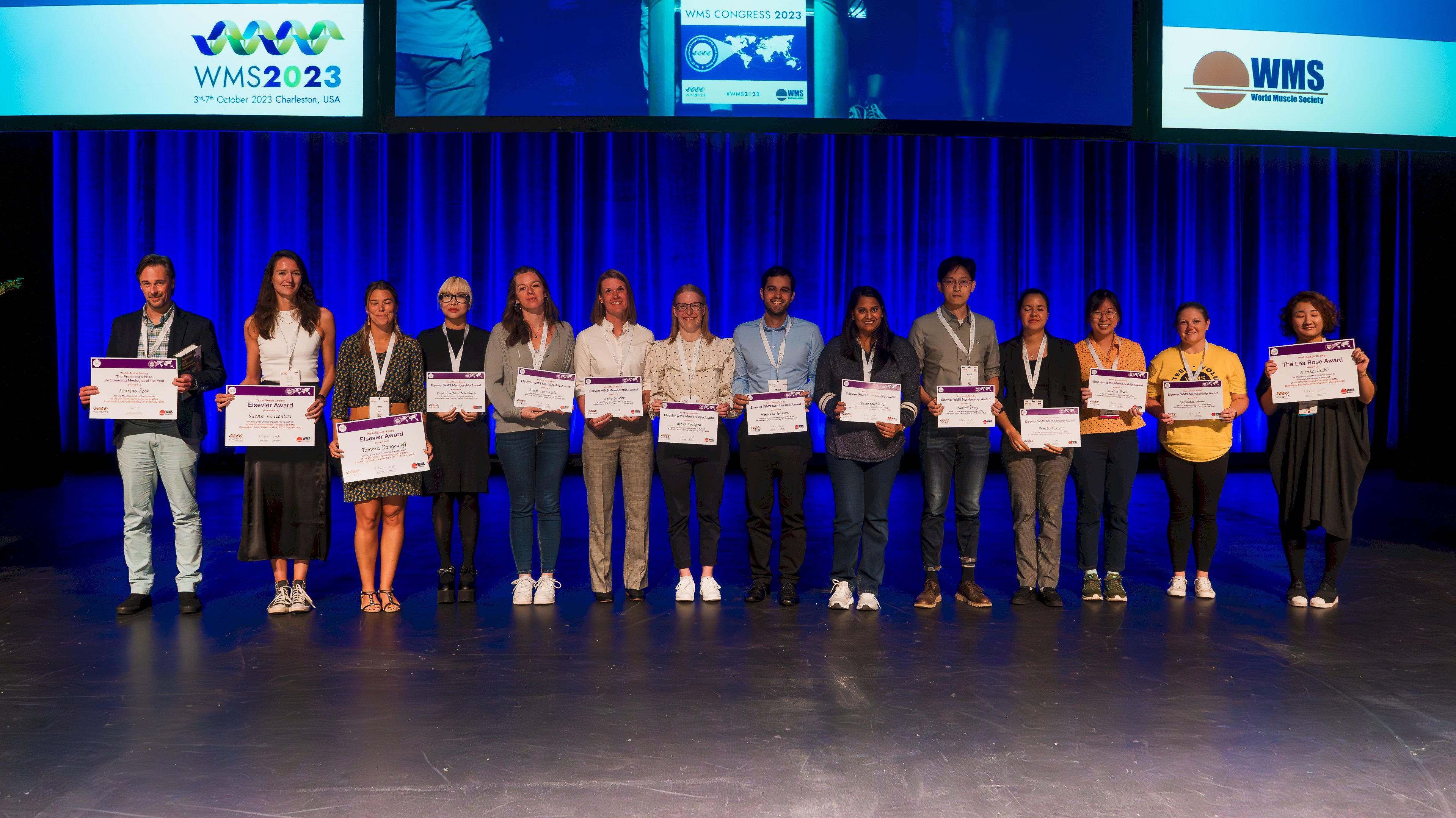 Prize winners on stage, each holding an A3 certificate in front of a blue backdrop