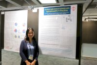 Dr Chan is dressed in a light blue top and dark blue cardigan. She is wearing an event lanyard and standing in front of a poster presentation board. 