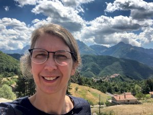 Dr Voermans stands outside smiling at the camera. The background is mountainous and lush. There are fluffy white clouds in the blue sky.
