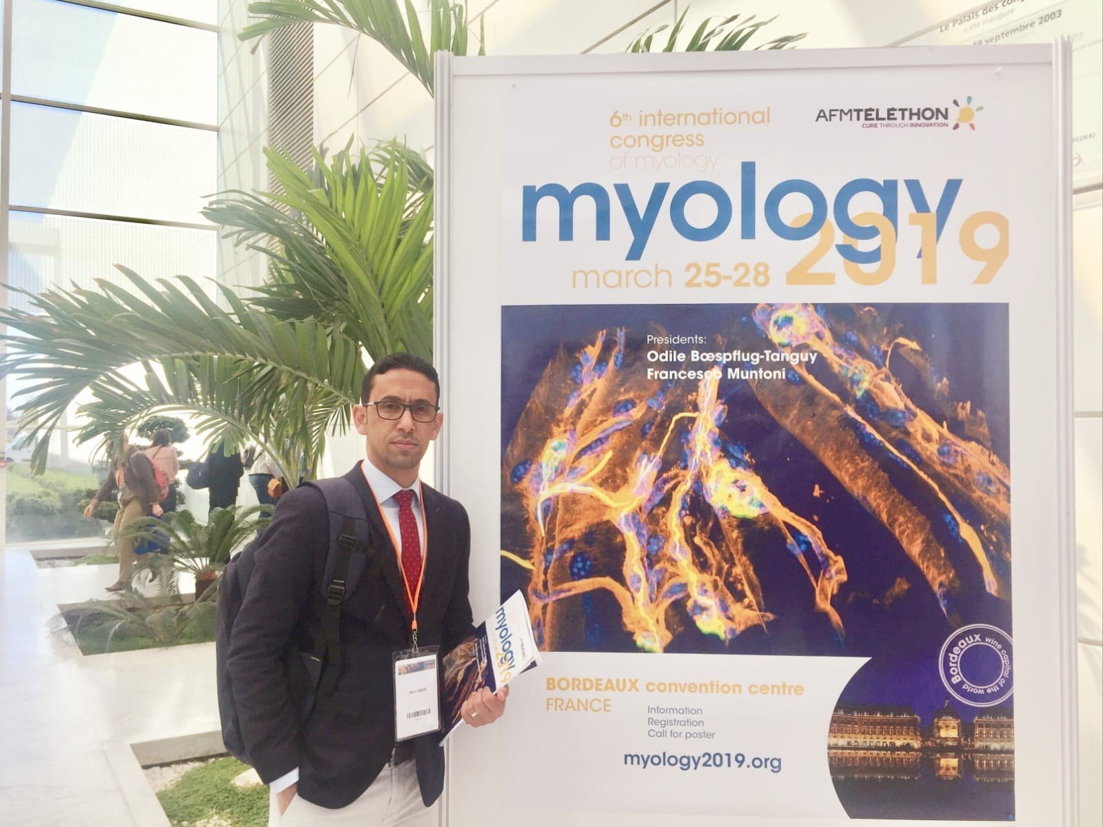 Professor Saleh Salman Omairi stands in front of a banner advertising Mylogy 2019. He is in the brightly lit atrium of a building with large palm trees in the background.