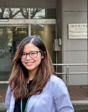 Genevieve is standing in front of a building and smiling at the camera. She has long dark hair and wears glasses and a white lab coat