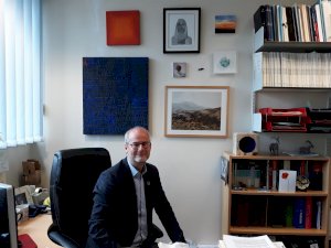Dr Volker Straub sits at his desk in his office at the University of Newcastle. He is wearing a dark suit with a light coloured shirt. On the wall behind him are various artworks. To the right, there are bookshelves packed with files and journals. 