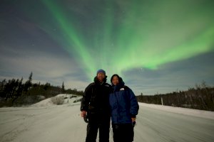 Dr Chan and her husband Mike stand in the foreground. The Ground is covered in snow. The sky is illuminated with the fluorescent glow of the Northern Lights. 