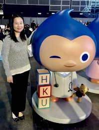 Dr Chan wears a grey jumper and black trousers. She is standing next to a model of a cartoon doctor with blue hair
